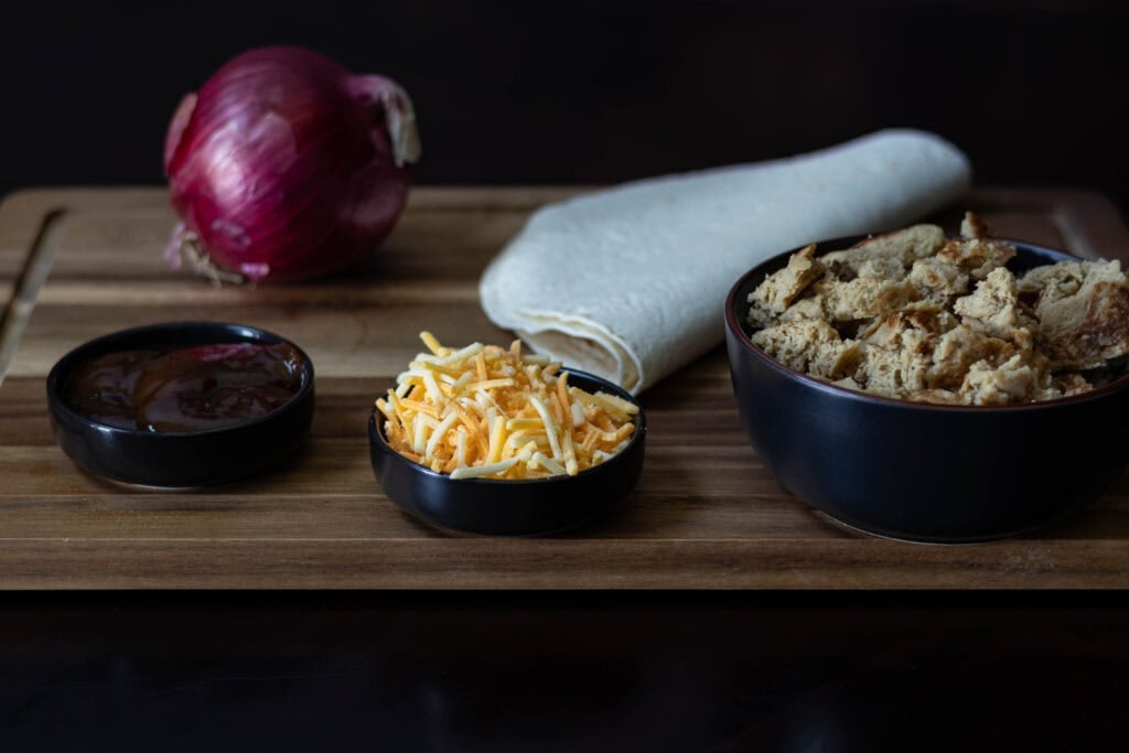 Ingredients for Shredded BBQ Seitan Quesadilla - BBQ Sauce, Vegan Cheese, red onion, tortillas, shredded seitan, sitting on a wooden cutting board.