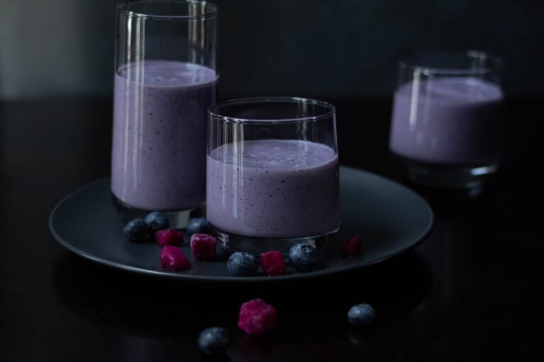 Three glasses with blueberry dragonfruit smoothie, sitting a grey plate, with a few pieces of blueberries and dragonfruit on the table.