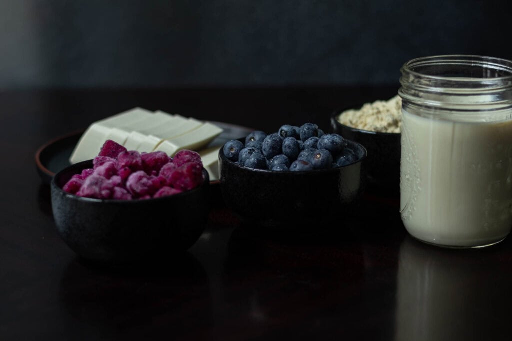 Ingredients for the blueberry dragonfruit smoothie sitting a table - silken tofu, protein powder, soy milk, dragonfruit and blueberries.