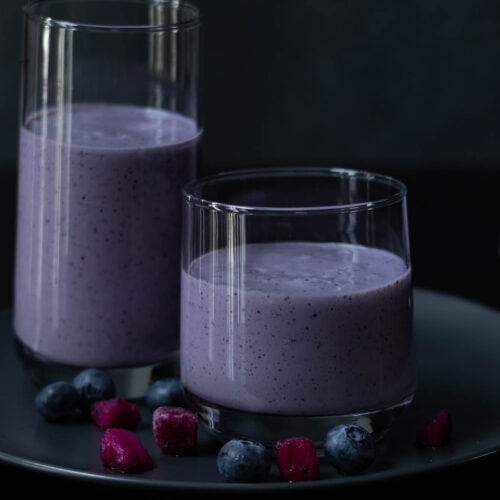 Two glasses with blueberry dragonfruit smoothie, sitting a grey plate, with a few pieces of blueberries and dragonfruit on the table.