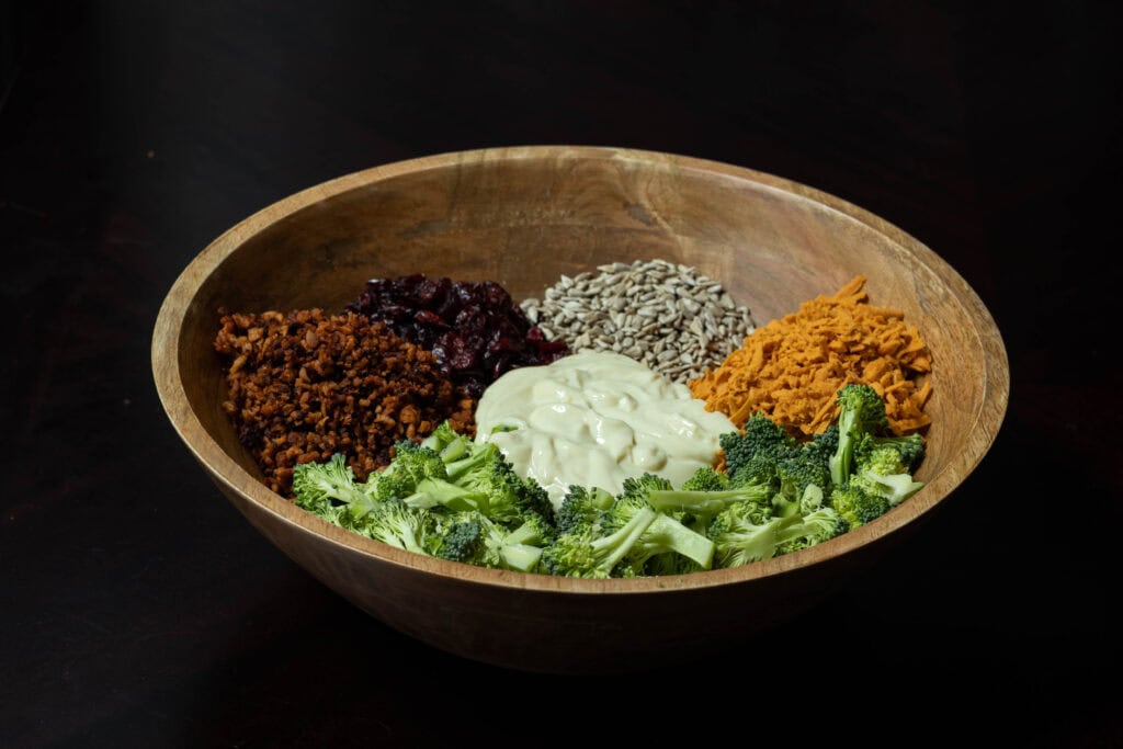 Large wooden bowl with broccoli salad ingredients - vegan bacon bits, broccoli florets, dressing, vegan shredded cheddar cheese, sunflower seeds and craisins.