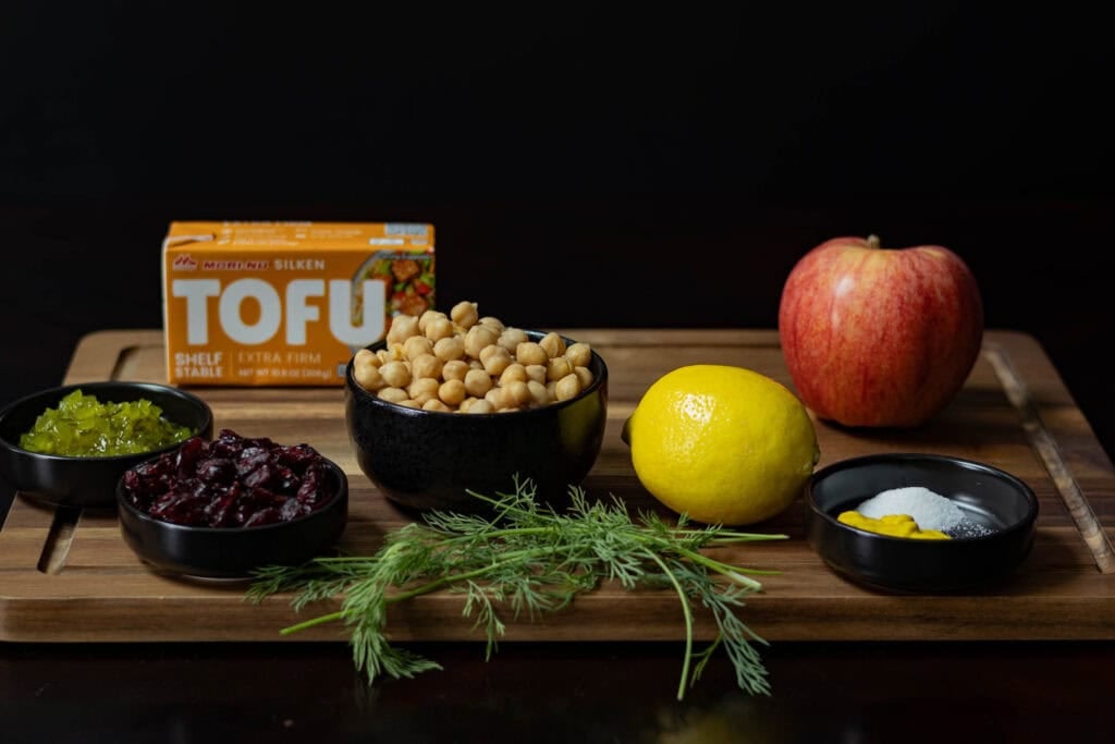 Ingredients for No-Mayo Chickpea Salad sitting on a wooden cutting board - fresh dill, craisins, relish, chickpeas, silken tofu, lemon, apple and spices.