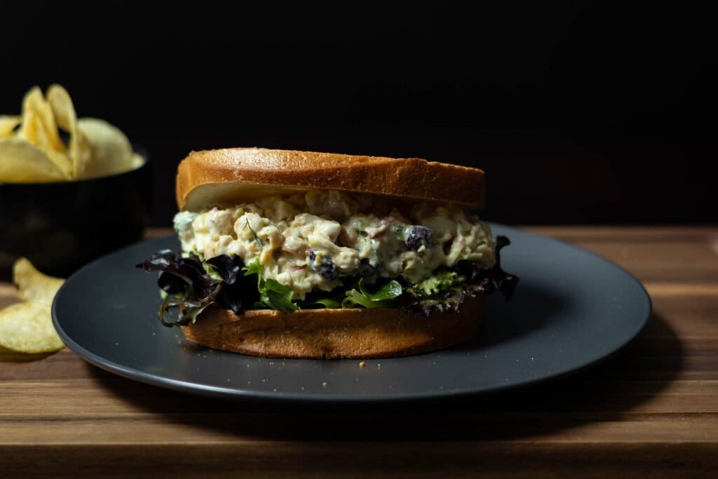 Chickpea Salad Sandwich with fresh mixed greens sitting on a grey plate, placed on a wooden cutting board with a bowl of chips in the background.