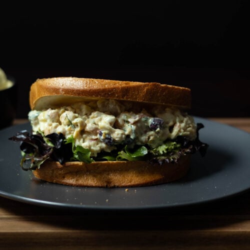 Chickpea Salad Sandwich with fresh mixed greens sitting on a grey plate, placed on a wooden cutting board with a bowl of chips in the background.