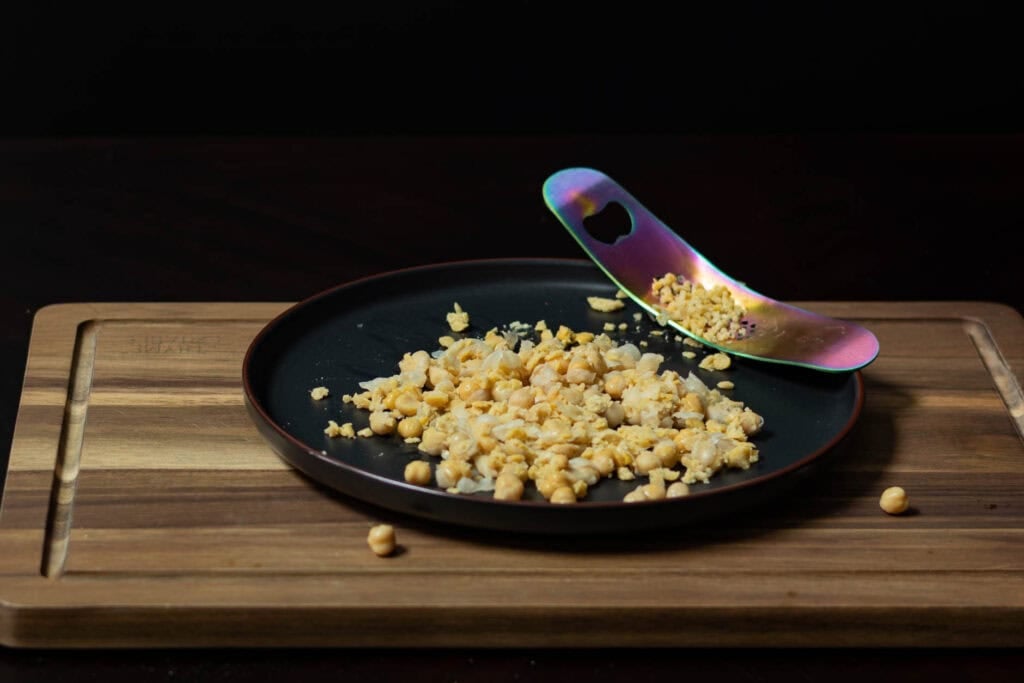 A black plate of smashed chickpeas sitting on a wooden cutting board.