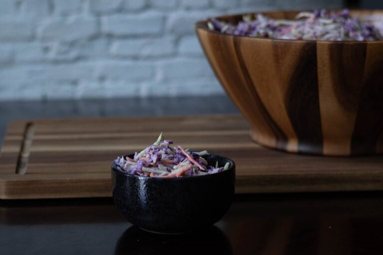 Small bowl of high protein cole slaw with a large wooden bowl of cole slaw in the background sitting on a wooden cutting board.