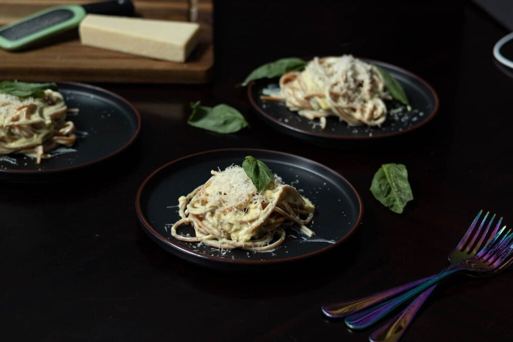 Three black plates with high protein alfredo garnished with basil leaves and a wooden cutting board with a block of vegan parmesan cheese and grater in the background.