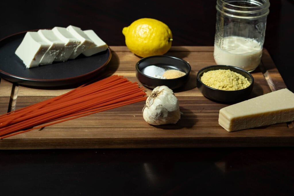 High Protein Alfredo ingredients sitting on a wooden cutting board - silken tofu, red lentil pasta, nutritional yeast, garlic, vegan parmesan cheese, soy milk, lemon and spices.