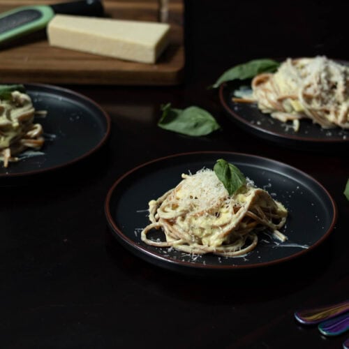 Three black plates with high protein alfredo garnished with basil leaves and a wooden cutting board with a block of vegan parmesan cheese and grater in the background.