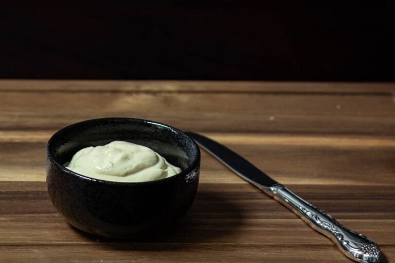 Small black bowl of high protein mayo with a knife to the side of it, sitting on a wooden cutting board.