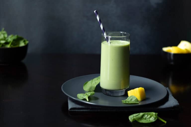 Green smoothie in a tall glass on a dark plate, with a bowl of spinach and a bowl of pineapple in the background.