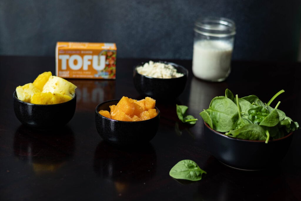 Smoothie ingredients on a table - silken tofu, pineapple, passionfruit, spinach, protein powder and soy milk.