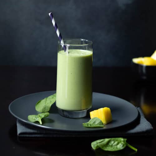 Green smoothie in a tall glass on a dark plate, with a bowl of pineapple in the background.