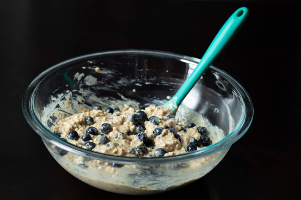 Mixed blueberry muffin batter in a large glass bowl with a blue spatula sitting on a dark table.