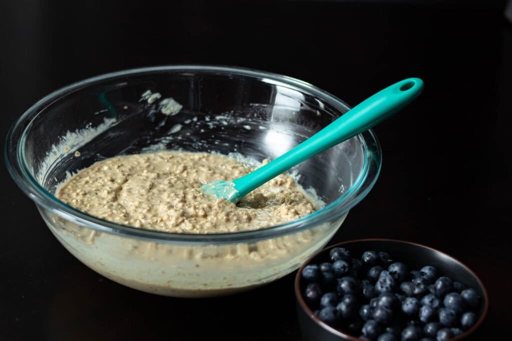 Large glass bowl of muffin batter with a smaller bowl of blueberries to the side.