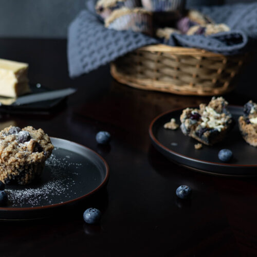 A small black plate with a blueberry muffin dusted with powder sugar, a small black plate with a muffin cut in half and buttered, a stick of butter and knife, a basket of muffins in the background, and a few blueberries on the table.