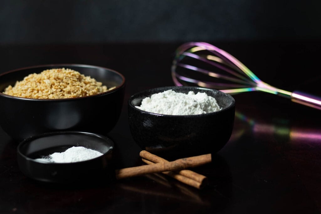 Dry ingredients for the muffins sitting on the table in black bowls next to a whisk - TVP, flour, cinnamon sticks, baking soda, salt, and baking soda.
