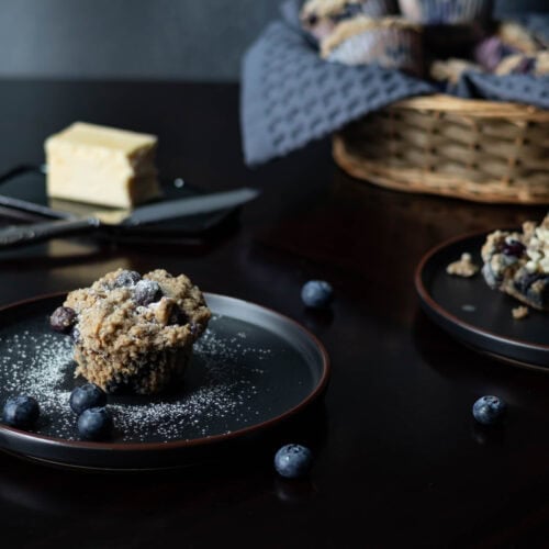 A small black plate with a blueberry muffin dusted with powder sugar, a small black plate with a muffin cut in half and buttered, a stick of butter and knife, a basket of muffins in the background, and a few blueberries on the table.