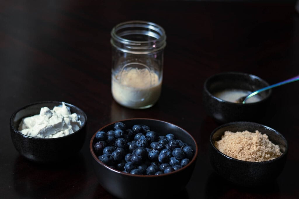 Wet ingredients for the muffins in bowls sitting on a dark table - vegan greek yogurt, blueberries, soy milk, flax egg and brown sugar.