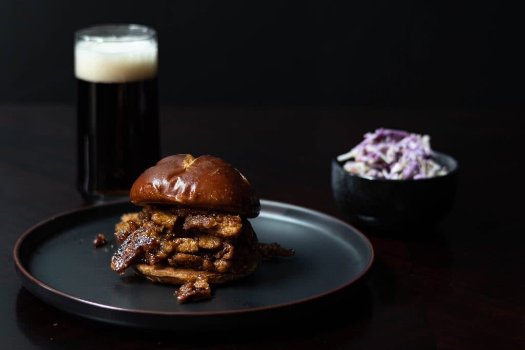 Shredded BBQ Seitan on a toasted pretzel bun sitting a round black plate, glass of root beer and bowl of cole slaw in the background.