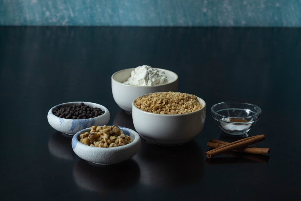 Dry ingredients for banana nut muffins in small bowls sitting on a table - mini chocolate chips, chopped walnuts, flour, TVP, cinnamon, salt, baking soda and baking powder.