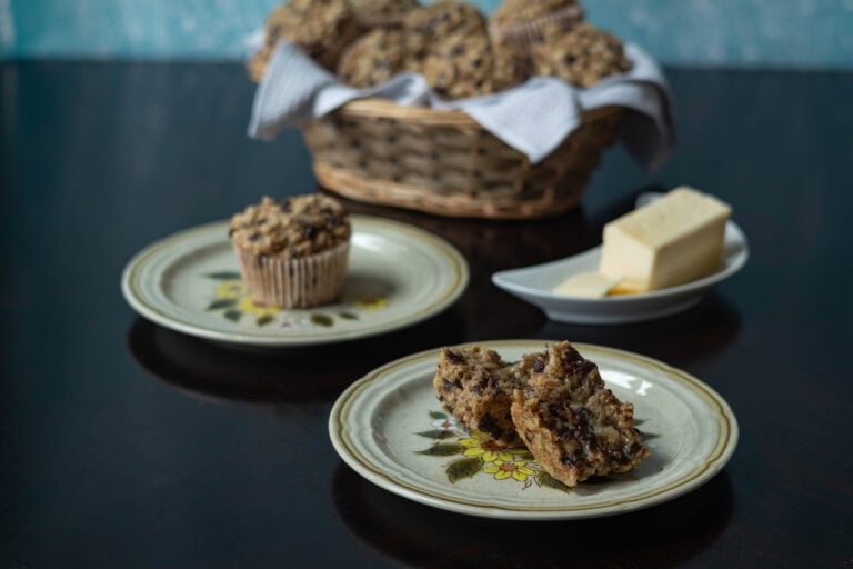 Cute little flower plates with banana nut muffins, with a large basket of muffins in the background and a crescent moon shaped plate of vegan butter to the side.