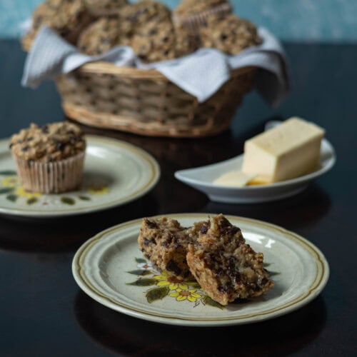 Cute little flower plates with banana nut muffins, with a large basket of muffins in the background and a crescent moon shaped plate of vegan butter to the side.