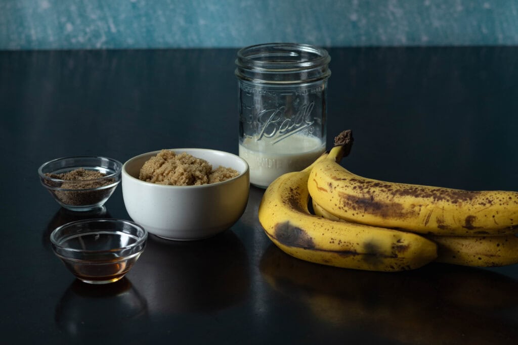Ingredients for banana nut muffins sitting on a wooden table - ground flax, vanilla extract, brown sugar, soy milk and bananas.