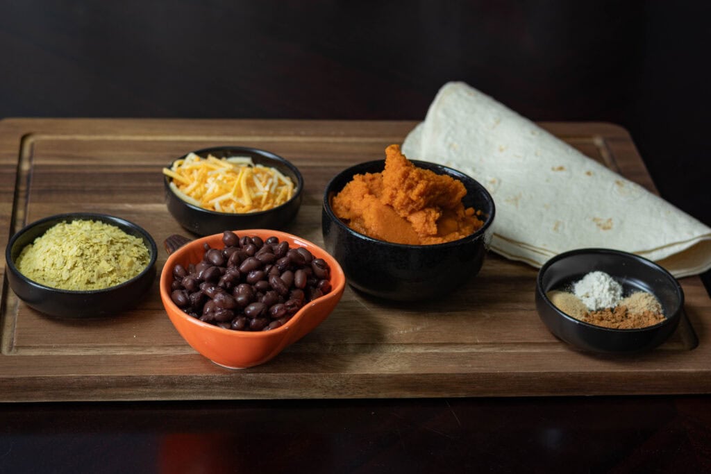 Ingredients for the Black Bean and Pumpkin Quesadilla sitting on a wooden cutting board - tortillas, spices, pumpkin puree, shredded vegan cheese, black beans, and nutritional yeast.