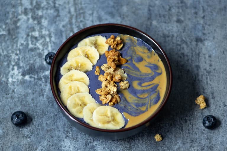 Blueberry smoothie bowl garnished with banana slices, granola and peanut butter swirl.