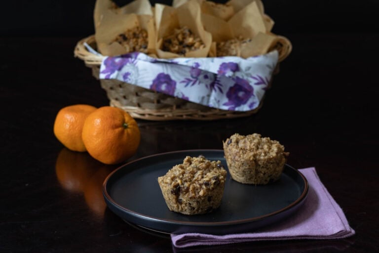 Black plate with two orange chocolate muffins. Two clementines, and a basket full of the remaining muffins in the background.