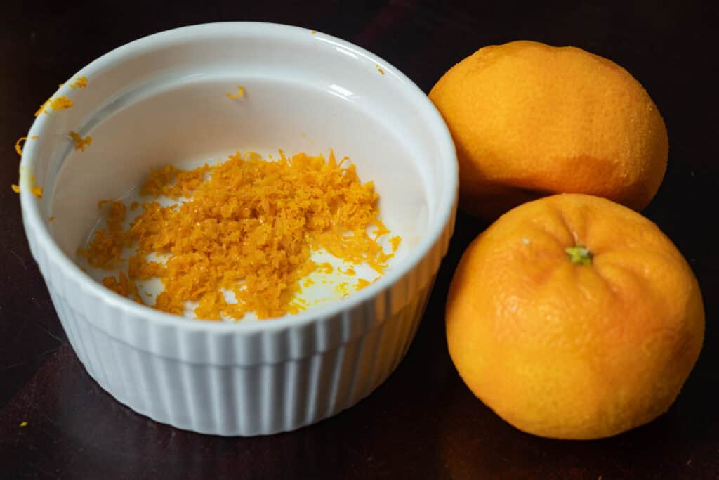 Two clementines sitting next to a white bowl of orange zest.