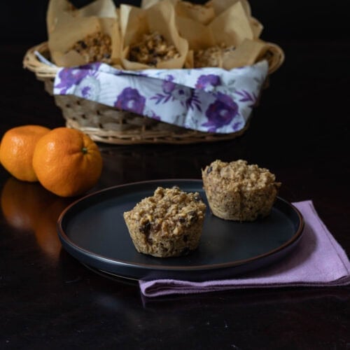Small black plate with two chocolate orange muffins in the front, with a basket of muffins in the background and two clementines to the side.