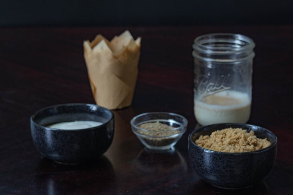 Muffin ingredients sitting on a wooden table - yogurt, ground flax, brown sugar, and soy milk.