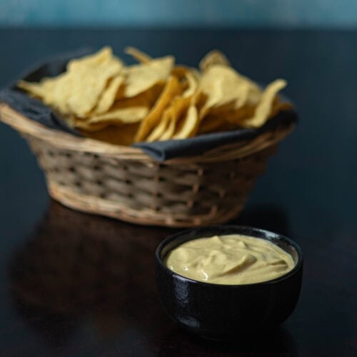 Bowl of high protein vegan cheese sauce with a basket of tortilla chips in the background.