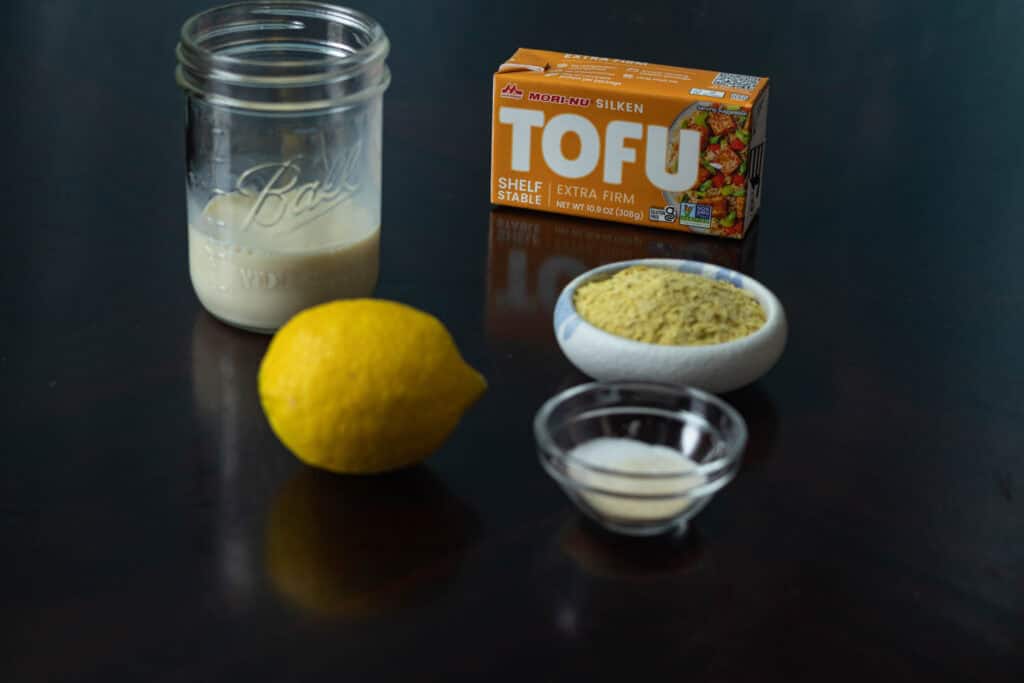 High Protein vegan cheese sauce ingredients sitting on a table - lemon, soy milk, silken tofu, nutritional yeast and spices.