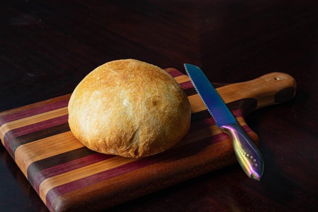 Freshly baked bread on wooden cutting board with serrated knife beside it.