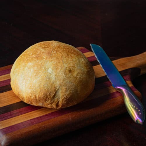 Freshly baked bread on wooden cutting board with serrated knife beside it.