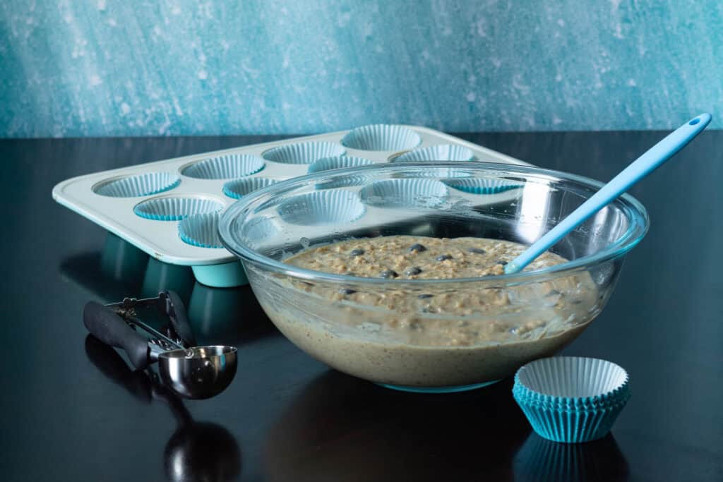 Large glass bowl with lemon blueberry muffin batter, lined muffin tray and ice cream scooper.