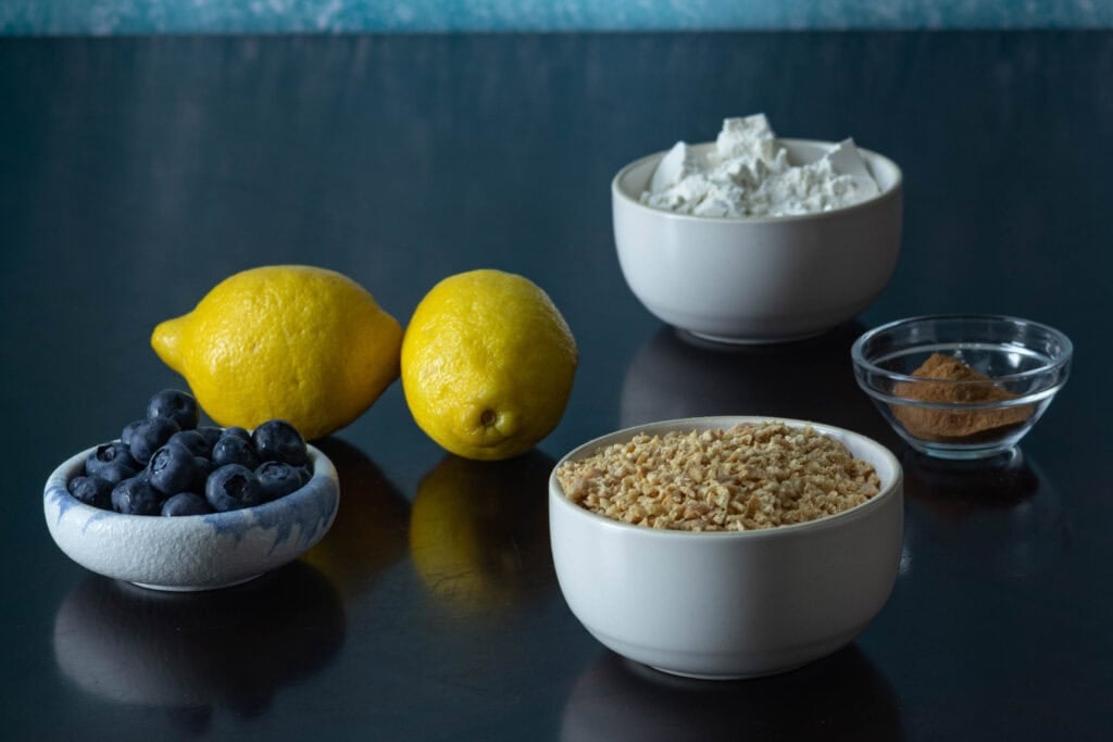 Muffin ingredients sitting on a wooden table - blueberries, lemons, flour, TVP and cinnamon, salt, baking soda, and baking powder.