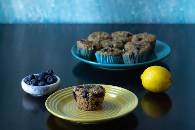 Small plate with a blueberry lemon muffin in front, larger plate with more muffins in the background, and blueberries and lemon to the side.