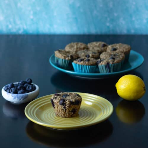 Small plate with a blueberry lemon muffin in front, larger plate with more muffins in the background, and blueberries and lemon to the side.