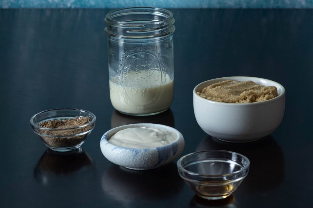 Muffin ingredients sitting on a wooden table - ground flax seed, vegan yogurt, vanilla extract, soy milk and brown sugar.