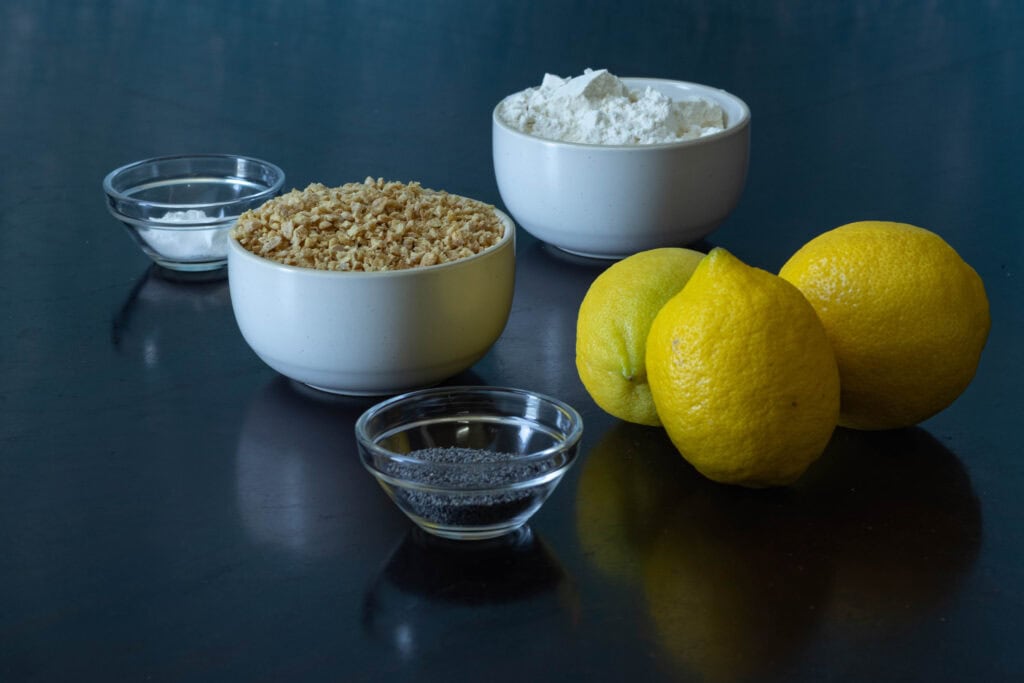 Ingredients for lemon poppyseed muffins sitting on a wooden table - flour, TVP, lemons, poppy seeds, salt, baking soda and baking powder.