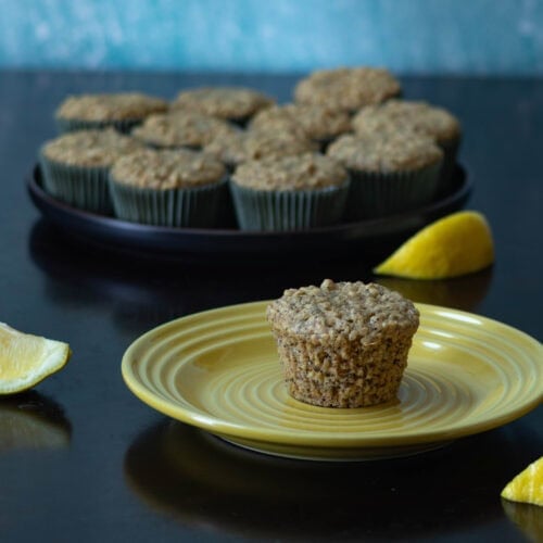 Lemon poppy seed muffin sitting on a small yellow plate, large black plate with more muffins in the background, and lemon slices.