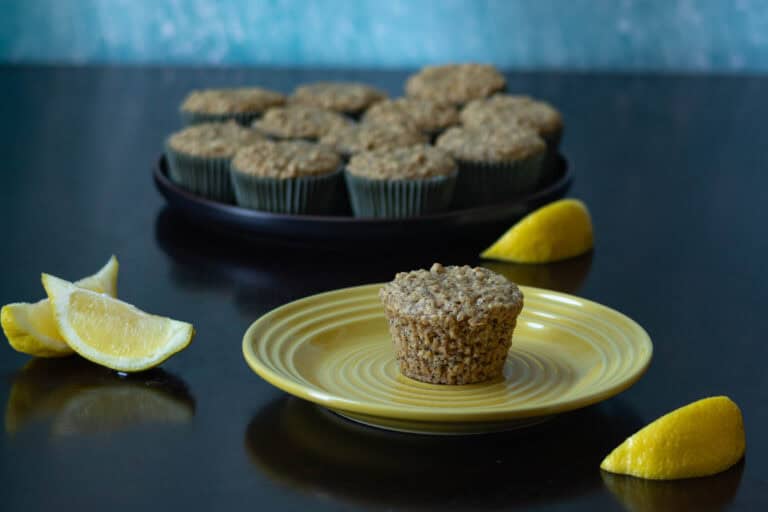 Lemon poppy seed muffin sitting on a small yellow plate, large black plate with more muffins in the background, and lemon slices.