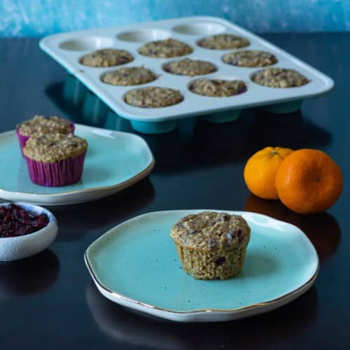 Orange craisin muffins sitting on light blue plates, muffin tray full of muffins in the background, and two clementines and a small bowl of craisins to the side.