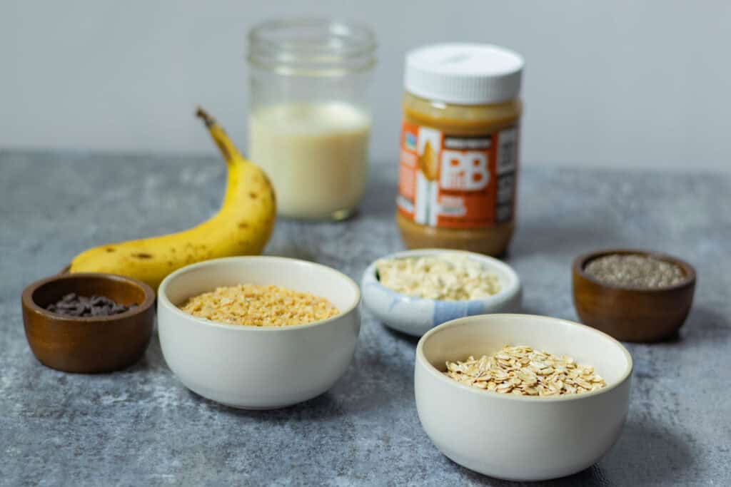 Overnight Oats ingredients sitting on a table - oats, TVP, protein powder, chia seeds, chocolate chips, soy milk, banana and peanut butter.