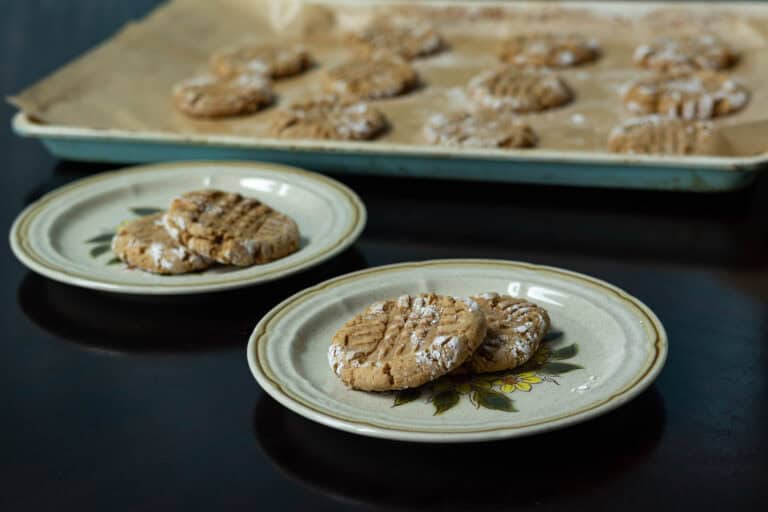 Two small plates with two peanut butter cookies each with a parchment paper-lined baking sheet with remaining cookies int he background.