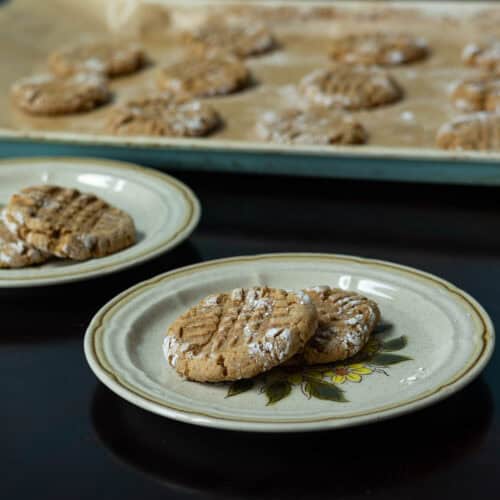 Two small plates with two peanut butter cookies each with a parchment paper-lined baking sheet with remaining cookies int he background.